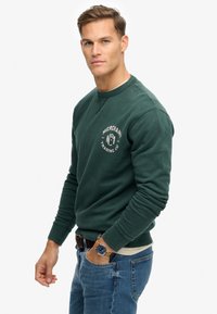 Sweat-shirt vert foncé à col rond, poignets côtelés et logo « Merchant Trading Co. » sur la poitrine, associé à un jean bleu et une montre.