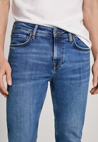 Jeans de mezclilla azul de corte ajustado, diseño de cinco bolsillos, con acentos de costura visibles y una cinturilla de altura media. La textura parece ligeramente desgastada.