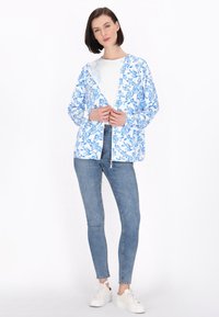 Jachetă ușoară cu model floral în alb și albastru, cu glugă, închidere cu șnur și mâneci lungi, asociată cu blugi albaștri și adidași.