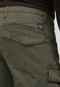 Un pantalon cargo vert olive avec une coupe slim, en tissu de coton, une grande poche arrière et une petite étiquette de marque à la taille.