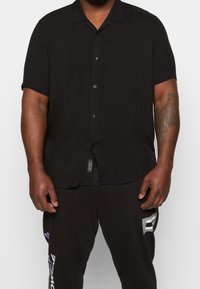 Zwart overhemd met korte mouwen van gladde stof, voorzien van een notch-kraag en een enkele pocket op de linkerkant van de borst.