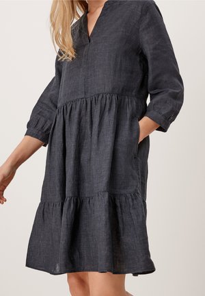 Kvinde iført en løs, mørkegrå hørkjole med opsamlede lag og trekvartlange ærmer, poserer med den ene hånd i lommen.