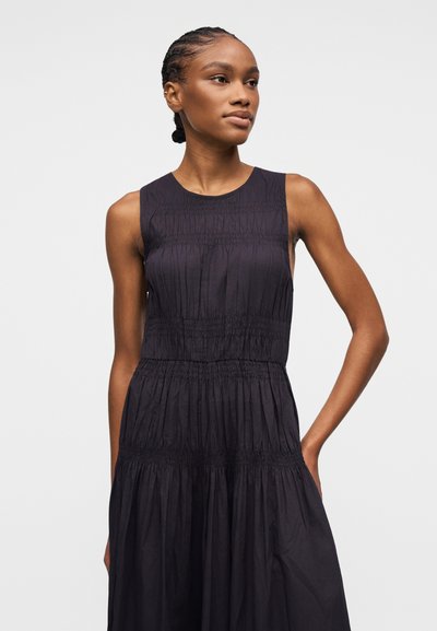 Femme aux cheveux tressés portant une robe noire sans manches avec des détails de tissu froncé, debout, une main sur la hanche.