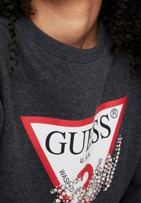 Sudadera gris con el logo de GUESS en rojo y blanco, que presenta adornos brillantes en la parte delantera en un patrón Curvo. Material de algodón, con cuello redondo.