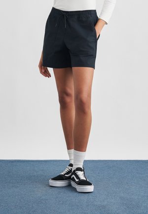 Person iført sorte shorts med snøre, hvid langærmet top, hvide sokker og sort-hvide sneakers, der står på blåt tæppe.
