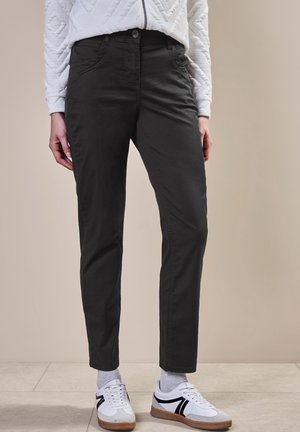 Osoba ubrana w czarne spodnie typu slim fit, białe trampki z czarnymi detalami, szare skarpety oraz białą pikowaną kurtkę stojąca na kafelkowanej podłodze.