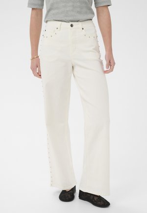 Frau trägt weiße, weit geschnittene Hosen mit Metallnieten an den Seiten, graues kurzärmeliges Oberteil, schwarze Spitzenschuhe und goldenen Schmuck an den Händen.