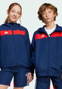 Deux individus portent des vestes de sport coordonnées bleu marine avec des rayures rouges, dotées de fermetures éclair et de poignets élastiques, assorties à des shorts bleu marine.