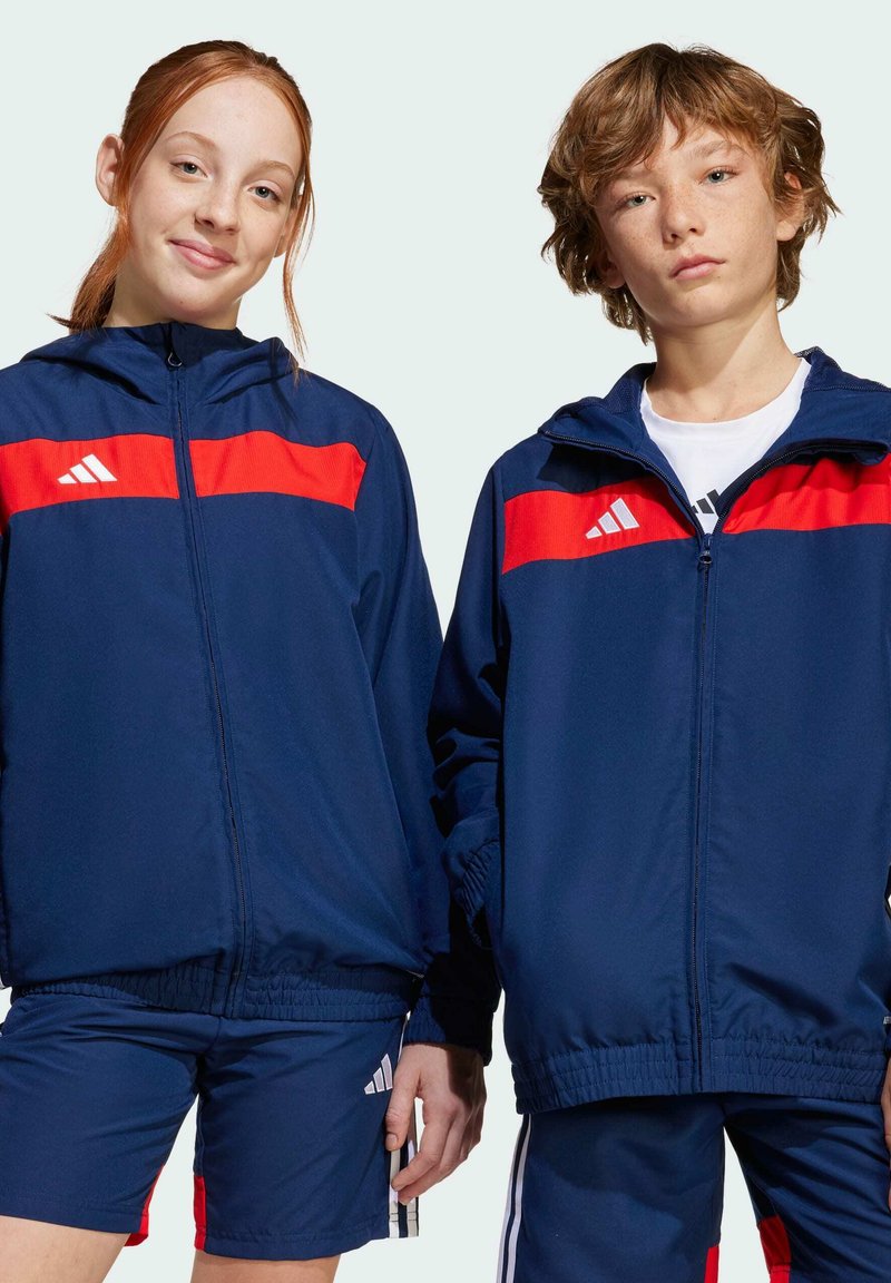Deux individus portent des vestes de sport coordonnées bleu marine avec des rayures rouges, dotées de fermetures éclair et de poignets élastiques, assorties à des shorts bleu marine.