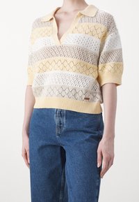 Pletený svetr s límcem a výstřihem do V, který má žluté, bílé a béžové diamantové vzory, zkrácený střih a krátké rukávy.