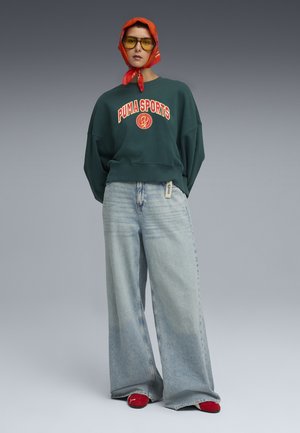 Grøn oversized sweatshirt med rød "PUMA SPORTS" logo, brede lyseblå jeans, røde sko og et rødt mønstret tørklæde. Teksturerede stofdetaljer er synlige.