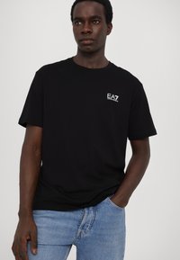 Svart t-skjorte med kort erm laget av et stoff med glatt tekstur. Har en liten hvit EA7-logo på venstre brystområde, kombinert med blå jeans.