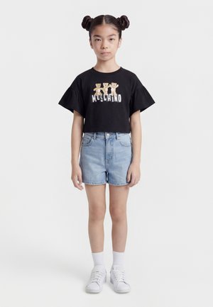 Ung pige med to knolde iført en sort t-shirt med bamseprint, lyse denimshorts, hvide sokker og hvide sneakers, stående mod en hvid baggrund.