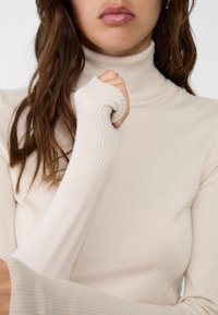 Beige coltrui met geribbeld textuur. Heeft lange mouwen met duimgaten en een aansluitende silhouet. Zachte, rekbare stof.