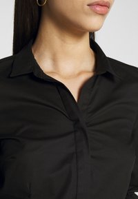 Femme portant une chemise noire à col et une boucle d'oreille anneau en or, avec cou, lèvres et cheveux noirs raides visibles sur un fond uni.