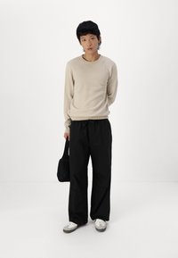 Beige striktrøje med ribbede manchetter, parret med løstsiddende sorte bukser. Modellen holder en lille sort taske og bærer hvide sneakers.
