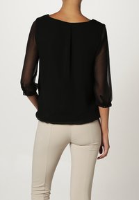 Femme portant un chemisier noir à manches trois-quarts transparentes et un pantalon beige ajusté, vue de dos sur un fond uni.