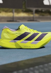 Scarpa da corsa giallo neon con strisce nere e dettaglio della suola "Lightstrike Pro" su una superficie blu della pista.