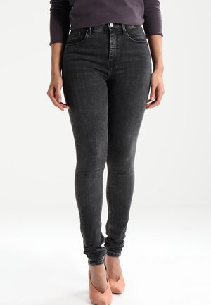 Kobieta ubrana w czarne, obcisłe jeansy z wysokim stanem, zestawione z brzoskwiniowymi szpilkami, stoi na tle jednolitego białego tła.