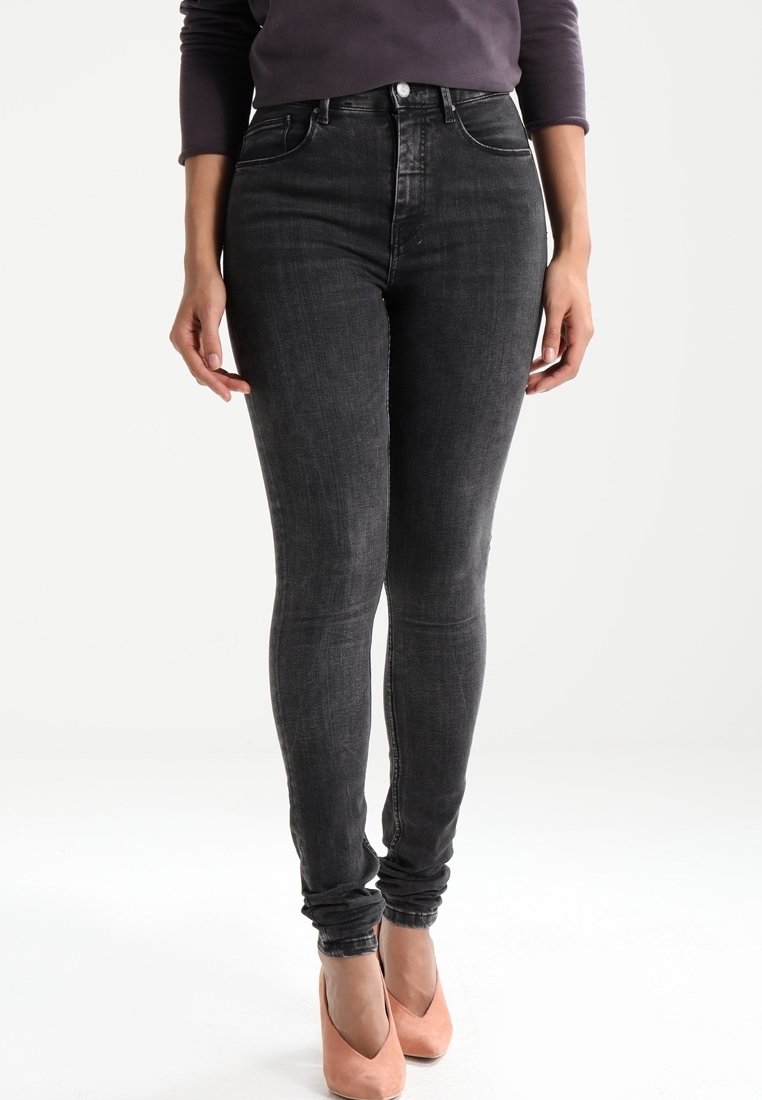 Femme portant un jean skinny noir taille haute associé à des escarpins pêche, debout devant un fond blanc uni.