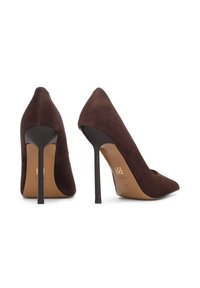 Tumši brūnas zamšādas augstpapēžu kurpes ar smailiem purngaliem un plāniem melniem stiletiem, attēlotas no aizmugures un sānu leņķa uz baltas virsmas.