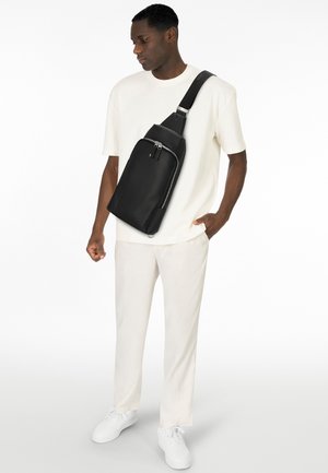 Borsa a tracolla in pelle nera con un design elegante e triangolare, tasca frontale con zip e tracolla regolabile. Indossata sopra una camicia bianca e pantaloni chiari.