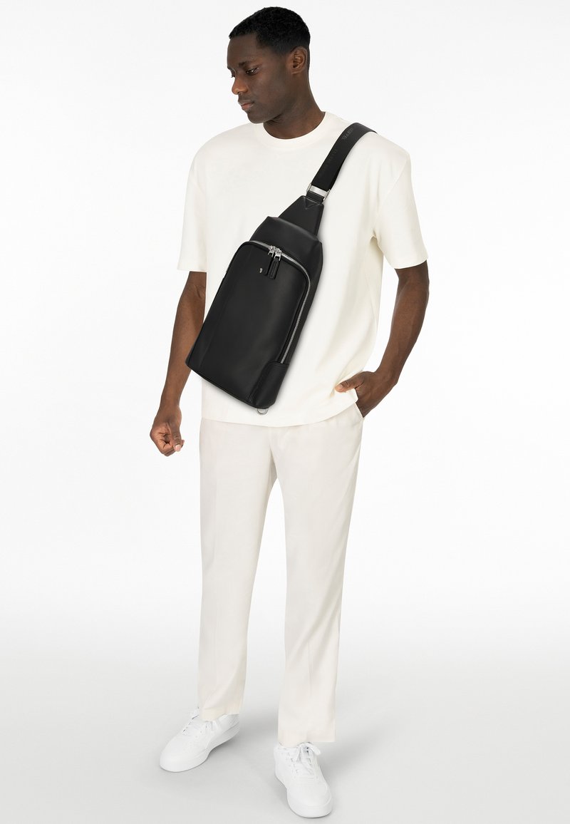 Bolso de cuero negro tipo bandolera con un diseño triangular elegante, bolsillo frontal con cremallera y correa ajustable. Llevado sobre una camiseta blanca y pantalones claros.
