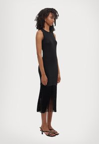 Svart ermeløs kjole med tettsittende bodice og frynset kant, parret med svarte sandaler med remmer. Glatt tekstur, knelengde silhuett.