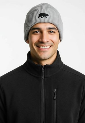Grijze ribgebreide muts met een zwarte beren silhouetlogo, gedragen met een zwarte fleecejas met ritssluiting zakken.