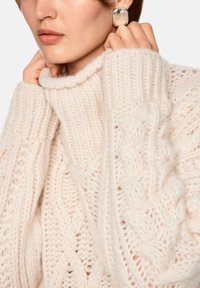 Grober Strickpullover in weichem Cremefarbton, mit geflochtenen und gerippten Mustern, hohem Kragen und dicken Bündchen. Sichtbarer silberner Statement-Ohrring.