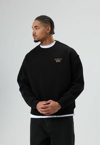 Zwarte sweatshirt met geribde halslijn, omgeslagen mouwen en een ontspannen pasvorm. Voorzien van een klein gouden geborduurd logo op de borst.