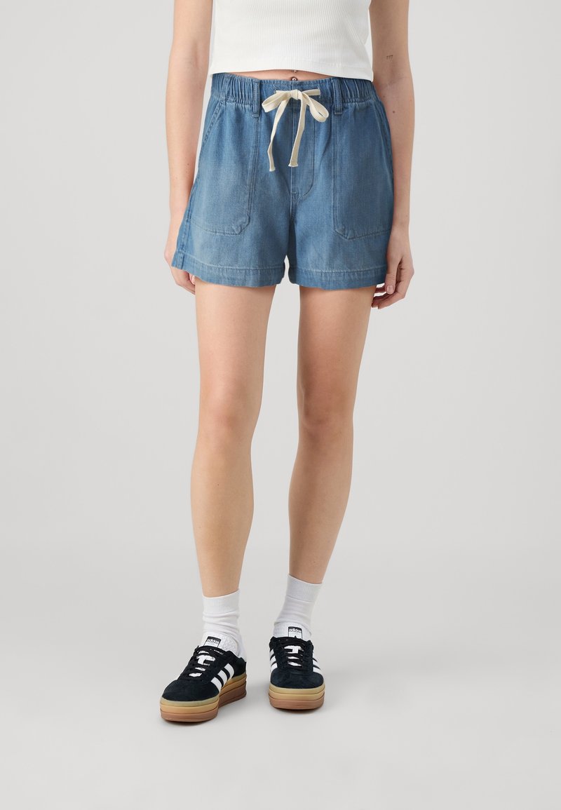 Personne portant un short en jean bleu clair avec taille élastique et cordon beige, un top court côtelé blanc, des chaussettes blanches et des baskets noires à rayures blanches.