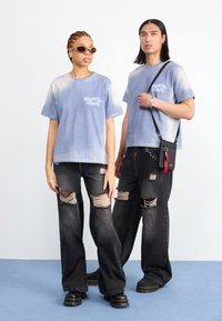 Du žmonės stovi šalia vienas kito dėvėdami derančius mėlynus t-shirt su dažų dėmėmis. Moteris derina savo marškinėlį su dėvėtomis juodomis džinsais ir akiniais nuo saulės. Vyras dėvi juodus džinsus su matomais įtrūkimais ir laiko mažą per petį nešamą maišelį. Abu dėvi juodus batus.