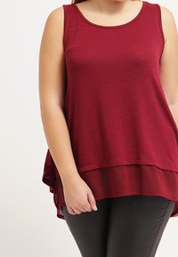 Burgunderfärgad ärmlös topp med rundad halsringning, lagerdesign och en kontrasterande genomskinlig nederkant. Tyget verkar mjukt med en subtil textur.