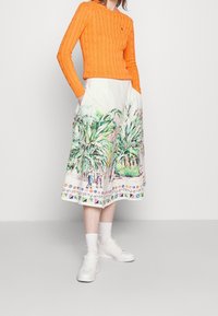 Nainen, joka käyttää kirkkaan oranssia palmikkoneuletta, yhdistettynä valkoiseen midi-hameeseen, jossa on värikkäitä trooppisia palmun ja hahmojen kuvioita.