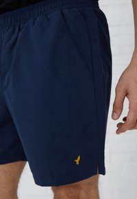 Námornícke šortky s elastickým pásom, bočnými vreckami a malým žltým logom vtáka pri okraji. Hladká, ľahká tkanina.