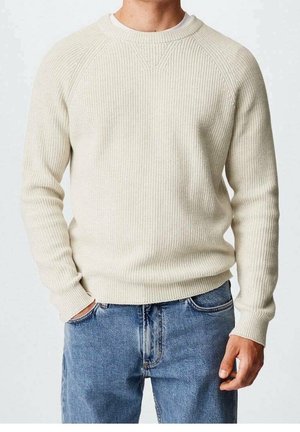 Homme portant un pull en tricot couleur crème et un jean bleu, avec une main dans la poche et des détails de couture visibles sur le pull.