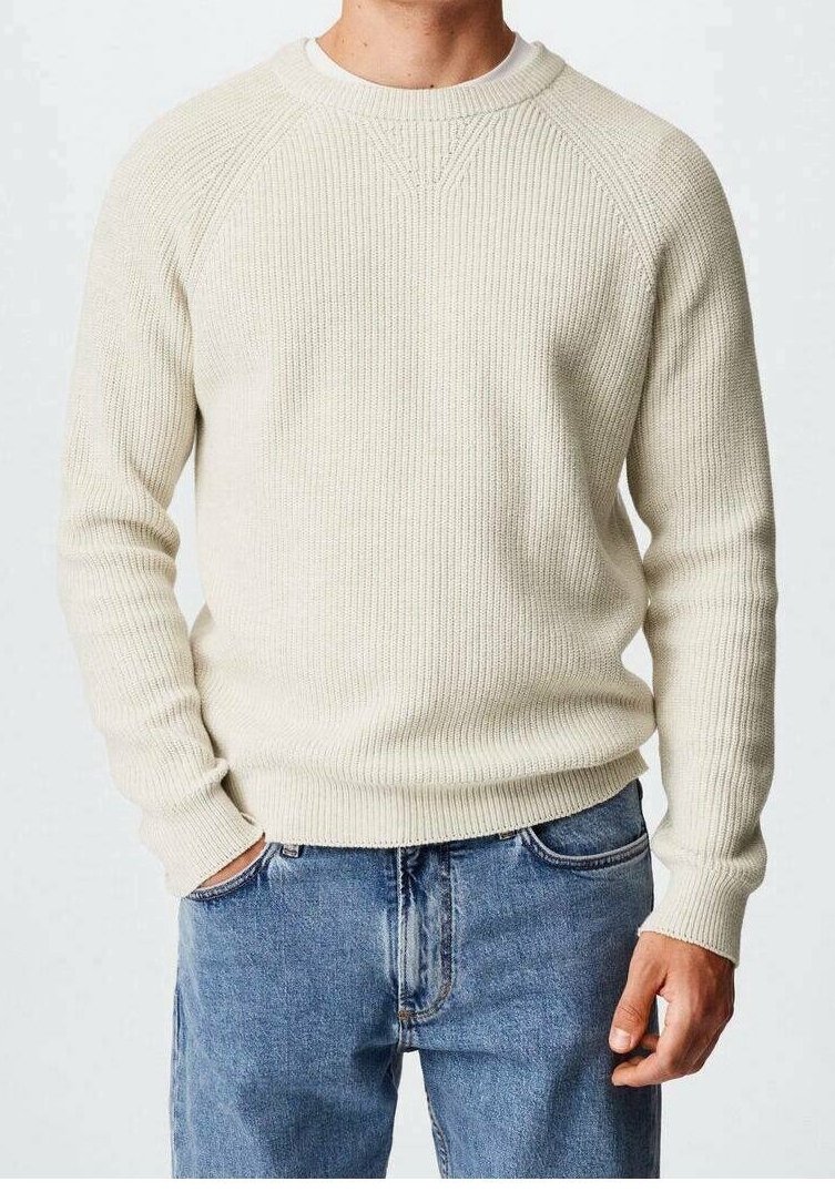 Homme portant un pull en tricot couleur crème et un jean bleu, avec une main dans la poche et des détails de couture visibles sur le pull.