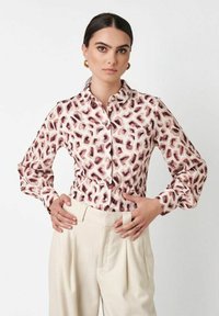 Frau mit dunklen Haaren, die goldene Kreolenohrringe, eine weiße Bluse mit braunem abstraktem Muster und hoch taillierte cremefarbene Hosen trägt, die Hände in die Hüften gestützt.