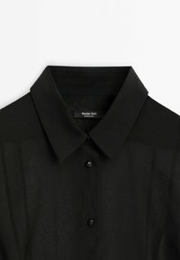 Musta paita klassisella kauluksella, jossa on läpinäkyvää kangasta ja nappikiinnitys, koristeena nelikulmaiset, mattapintaiset napit. Merkkilogo näkyvissä.