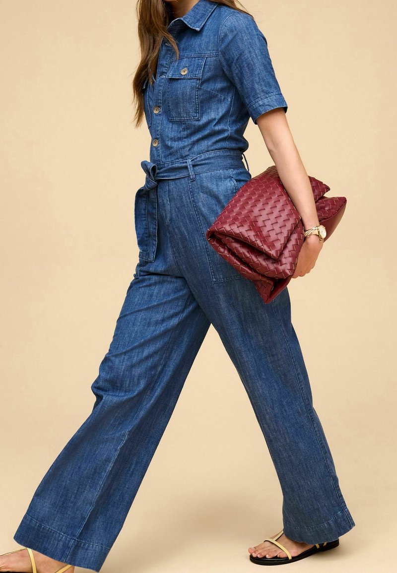 Femme en combinaison en denim bleu portant une grande pochette tissée rouge, une montre en or et des sandales plates beiges, marchant sur fond beige.