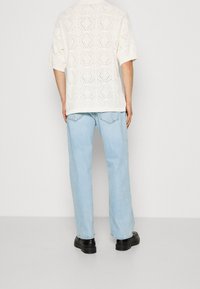 Witte gebreide top met korte mouwen en ruitpatronen, gecombineerd met lichtblauwe loszittende jeans en zwarte schoenen, weergegeven vanuit de achterkant.