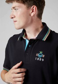 Polo noir avec des accents de col rayés multicolores, col intérieur à motifs et broderie "BOW 1859". Coupe décontractée et manches courtes.