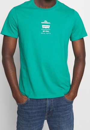 Homme portant un t-shirt à manches courtes Levi's bleu sarcelle avec logo blanc et un jean décontracté bleu, mains dans les poches, sur un fond uni.