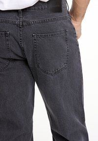Jeans de mezclilla negra con un ajuste relajado, que cuentan con bolsillos traseros y una etiqueta de marca de cuero en la cinturilla. La tela tiene una textura suave.