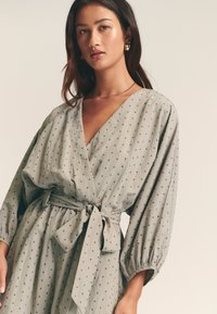 Lichtgrijze wikkeljurk met een diepe V-hals, geplooide mouwen en een gestrikte taille, met een subtiel patroon van kleine donkere stippen.