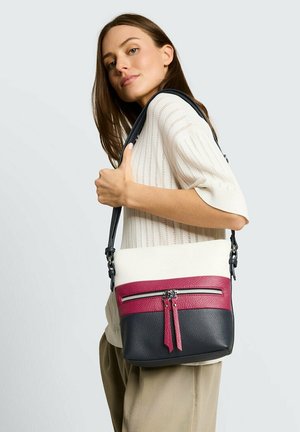 Frau in weißem Strickoberteil und beigen Hosen, die eine schwarze, weiße und pinke Leder-Umhängetasche mit Reißverschlussdetails trägt.