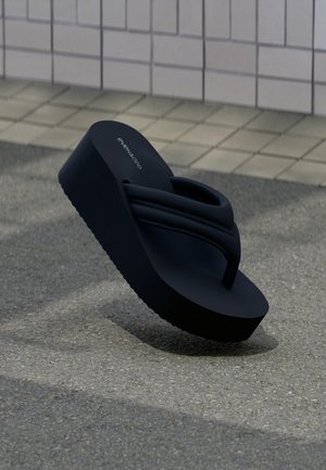 Schwarze Plateau-Zehensandale mit gepolsterten Riemen, schräg auf strukturiertem Boden vor einem gekachelten Wandhintergrund positioniert.