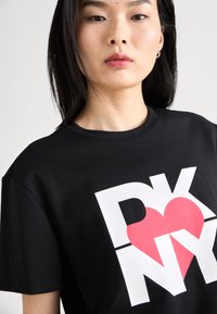 Svart bomull T-skjorte med en stor hvit "DKNY" logo med en rød hjerteform. Rund hals og korte ermer med en glatt tekstur.