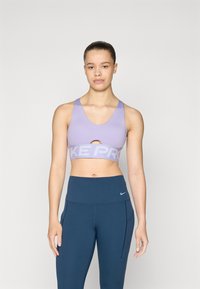 Vijolični športni modrček z izrezom v obliki obroča in širokim pasom, označenim z napisom "NIKE PRO". Pairs z temno modrimi visokimi legicami.